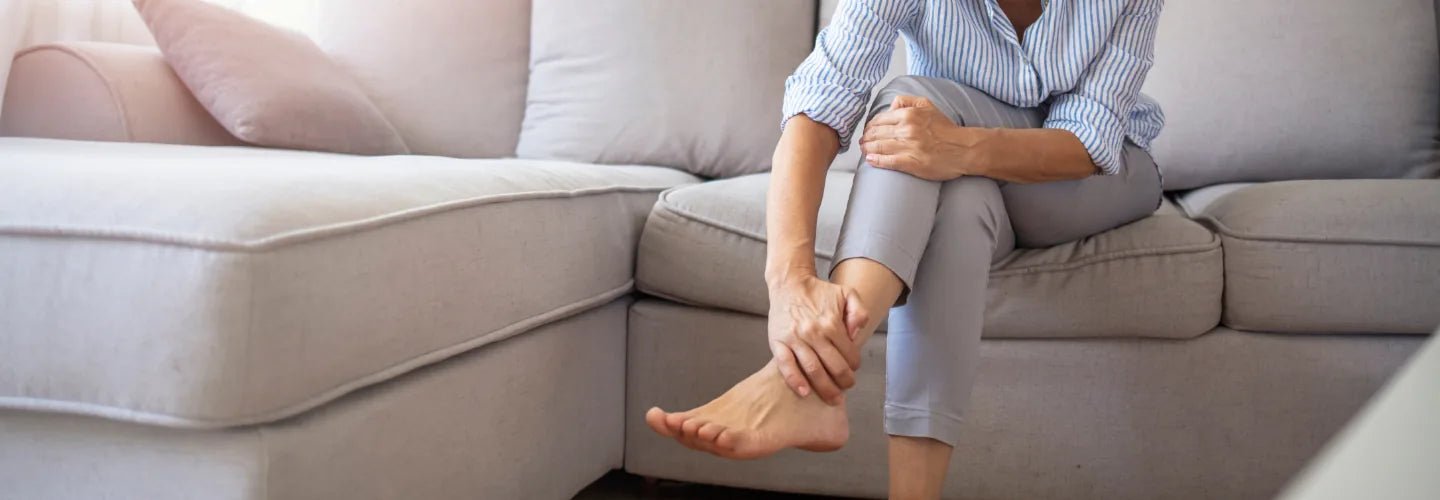 Ältere Frau sitzt zu Hause auf einem Sofa und berührt ihren Unterschenkel